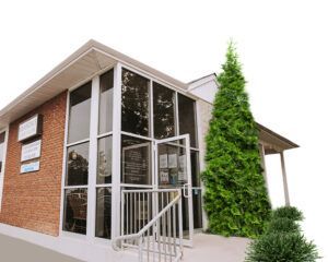 Brick building with large windows and open door. Tall evergreen tree next to the entrance.