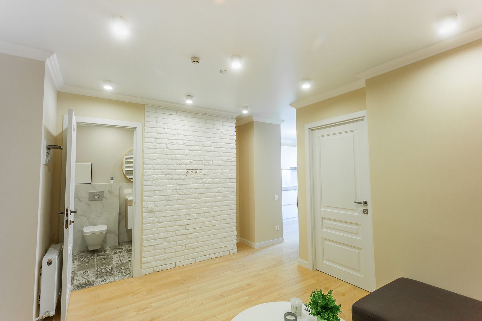 Interior hallway with access to a bathroom and other rooms. White brick wall, light wood floors, and recessed lighting.