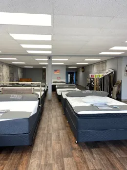 A showroom featuring rows of new mattresses on dark gray bed frames, arranged on a wood-look floor under ceiling lights.