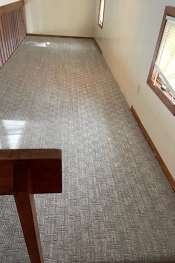 A light-filled hallway with patterned gray carpet, white walls, wood trim, and windows on the right side.