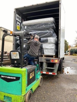 A worker stands on a green Mitsubishi forklift, loading a stack of mattresses into the back of a large semi-truck.