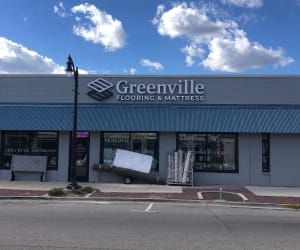A storefront for Greenville Flooring & Mattress, featuring a gray facade, blue awning, and large display windows.