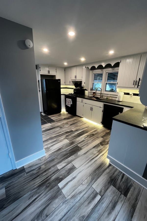 A modern kitchen with grey and white cabinetry, a black refrigerator, and grey wood-look flooring under recessed lighting.