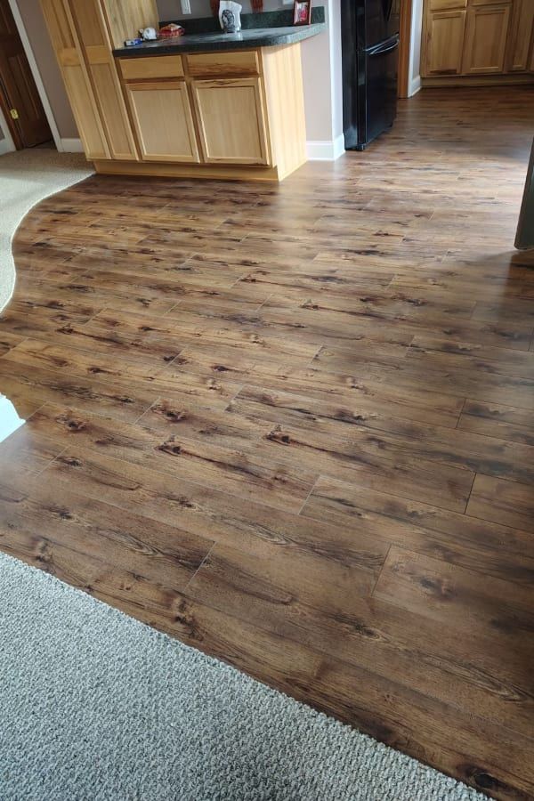 Transition between a grey carpet and dark brown wood-look vinyl plank flooring in a kitchen area with wooden cabinets.