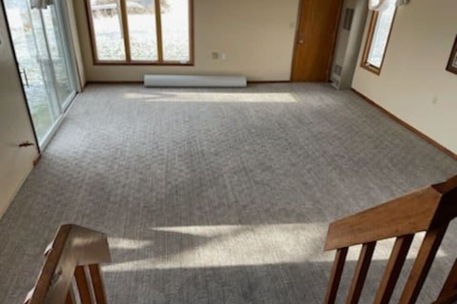 A high-angle view of a carpeted living room with light-colored carpet, large windows, a sliding door, and wooden stairs.