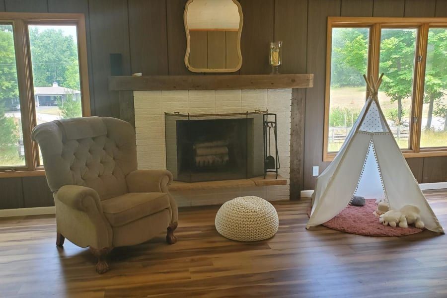 A room with wood walls and floors, a white brick fireplace, a tan wingback chair, a round knit pouf, and a small play tent.