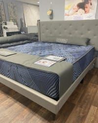 A tufted gray upholstered bed frame with a blue patterned mattress in a showroom with wood flooring.