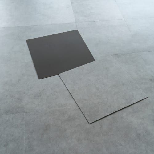 A dark square tile rests partially on top of a larger, light gray concrete-textured tile on a matching tiled floor.