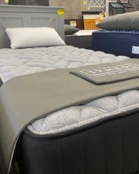 A gray bed frame with a textured white mattress and pillow, displaying a Symbol brand label on a gray runner.