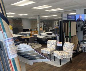 A showroom features patterned chairs, sample flooring displays, and several mattresses on pedestals under bright lighting.