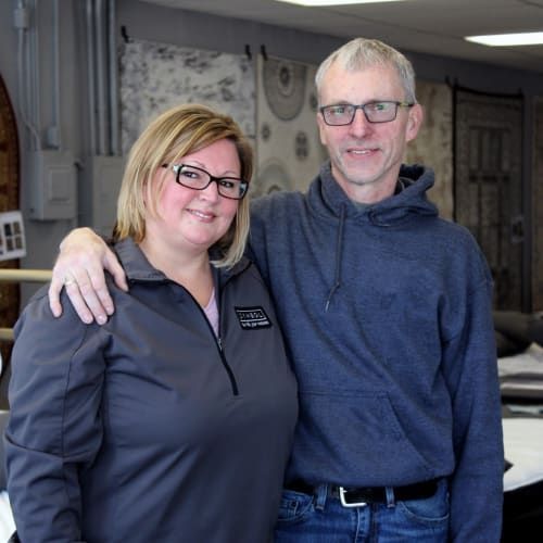 A smiling couple stands side-by-side in a store, with the man’s arm around the woman, both wearing glasses and casual wear.