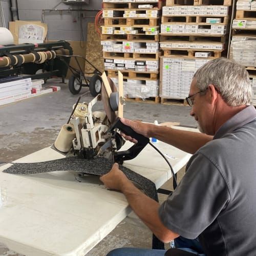 A person in a gray shirt uses a handheld industrial sewing machine to edge a piece of gray carpet on a folding table.