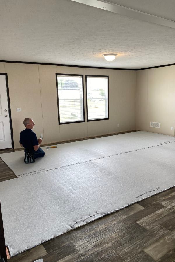A person kneeling on a floor covered in white underlayment, preparing to install new flooring in a room with two windows.