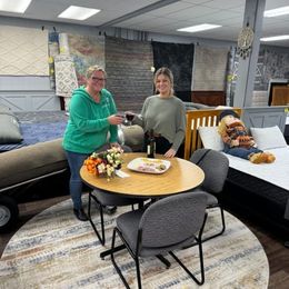 Two people toast with glasses while sitting at a small table inside a carpet and furniture showroom.