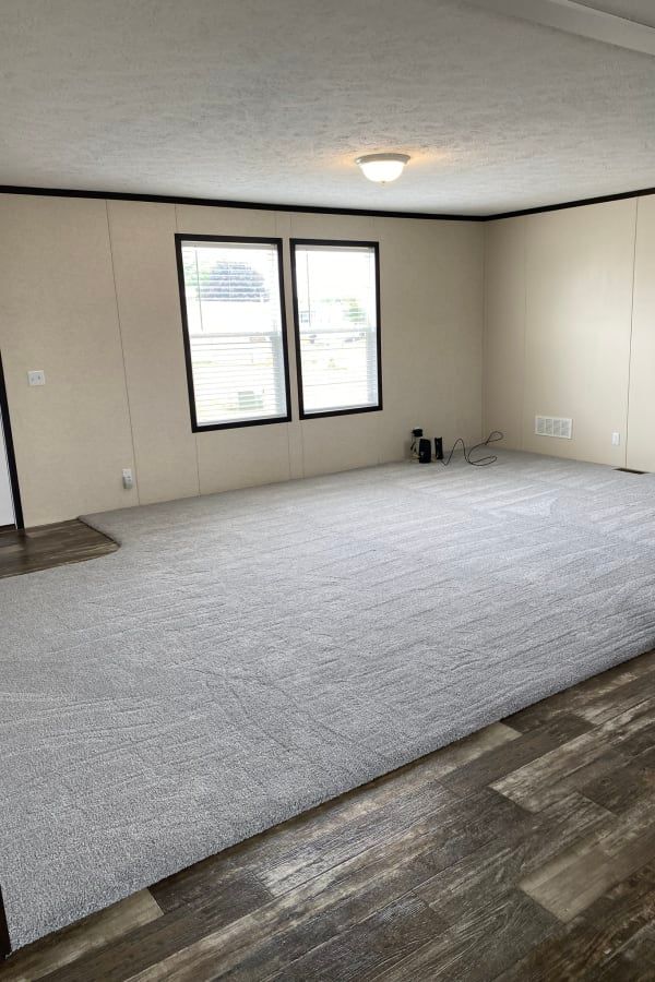 A spacious living area with light-colored walls, grey carpet, and wood-look flooring, featuring two windows.