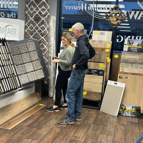 Two people stand in a flooring showroom, examining a display of carpet samples.