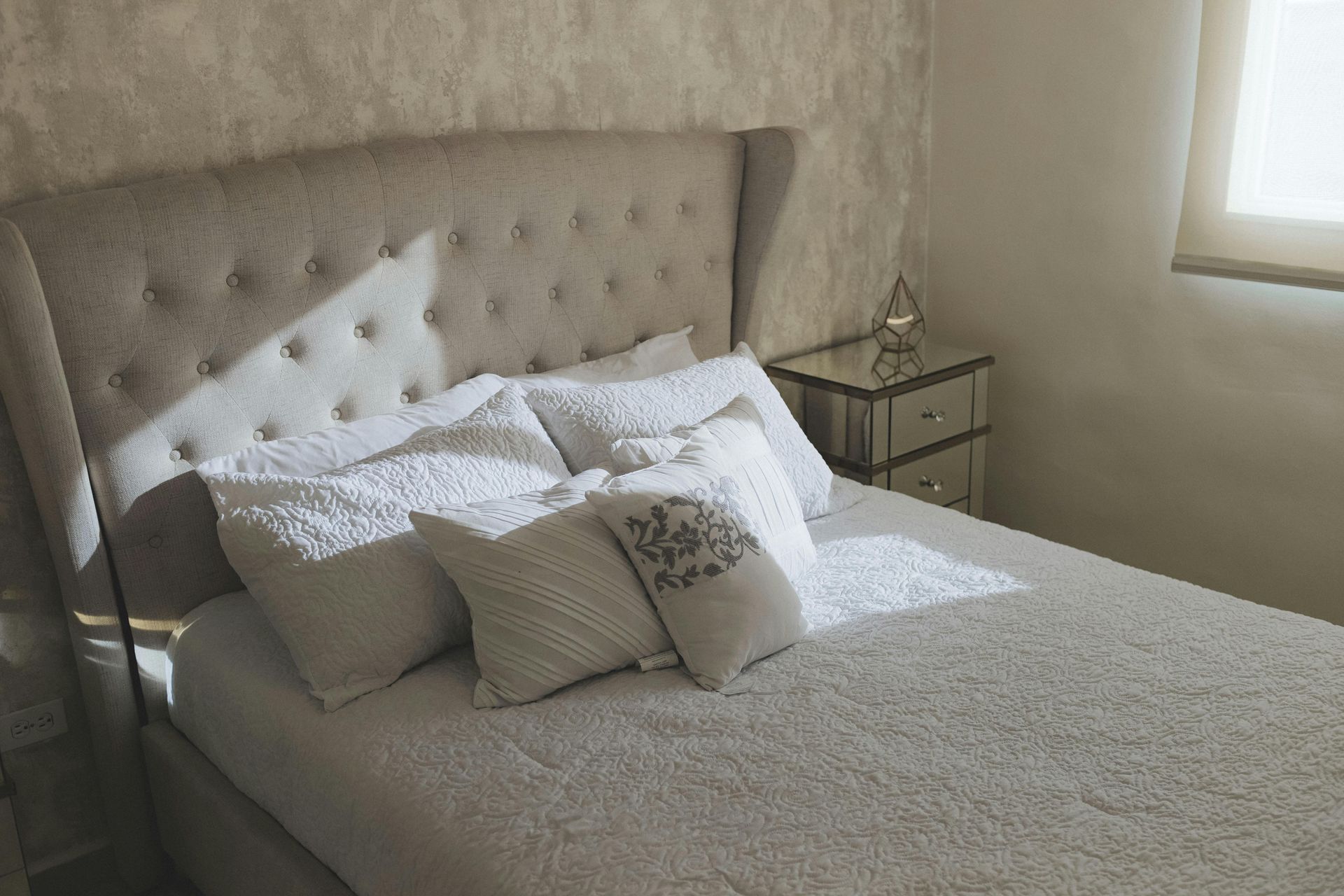 A tufted beige headboard and plush bedding in a neutral bedroom with a mirrored nightstand next to the bed.