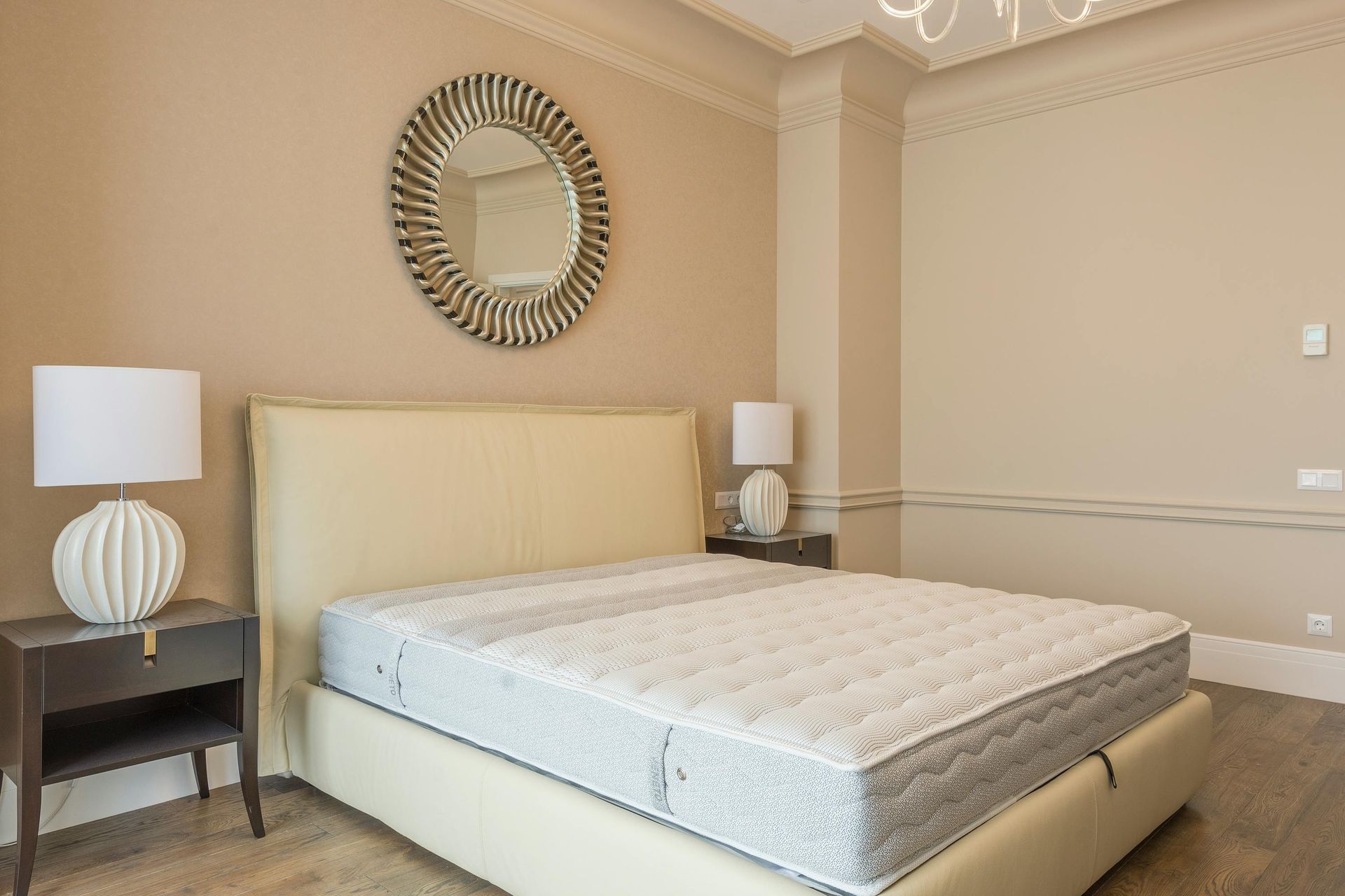A bedroom with a beige upholstered bed, matching nightstands with white lamps, and a sunburst mirror on the wall.