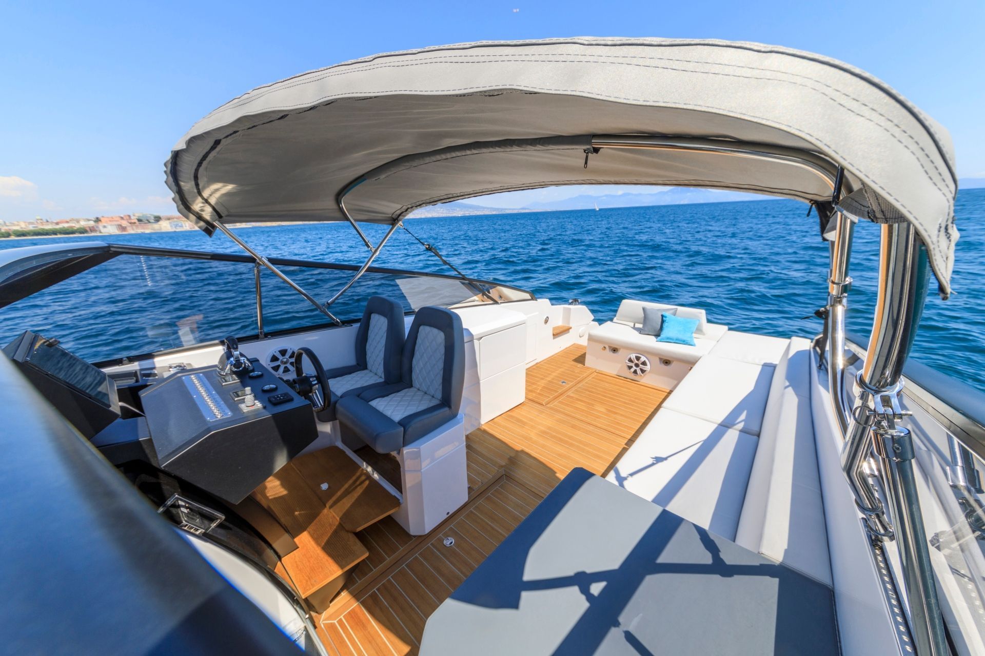 The open deck of a motorboat featuring a helm, seating, and a sun pad under a canopy, overlooking the open blue sea.