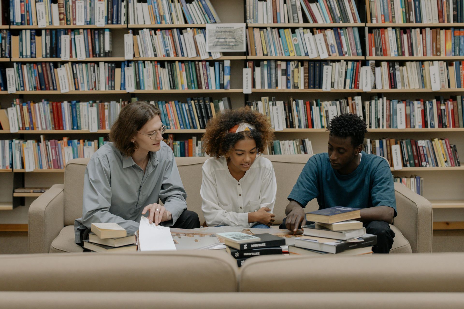Three people are sitting on a couch in a library looking at books.