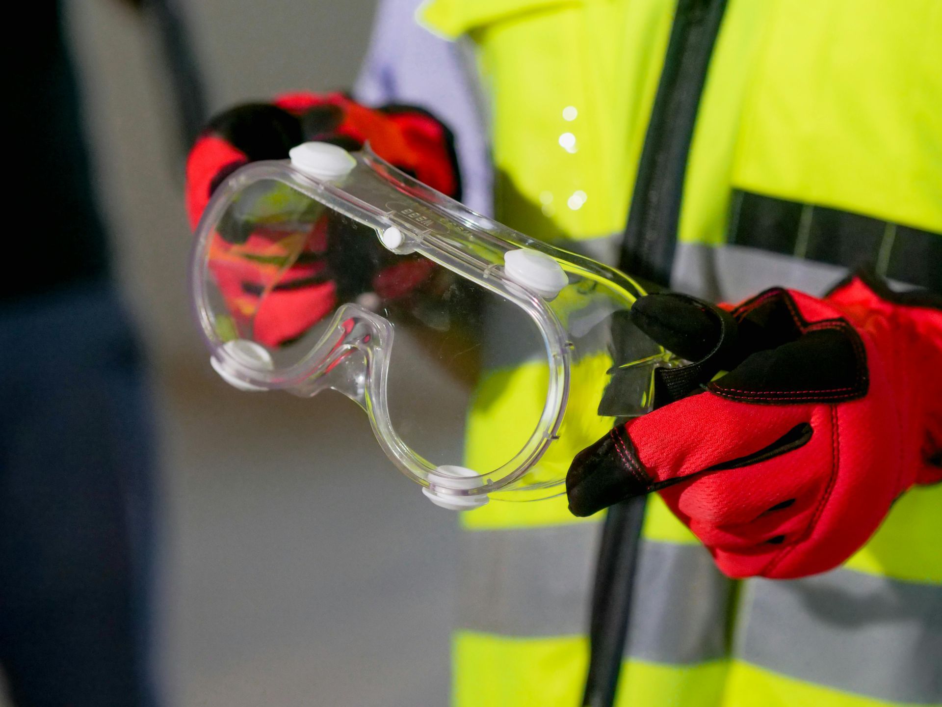 A person wearing a yellow vest and red gloves is holding a pair of safety goggles.