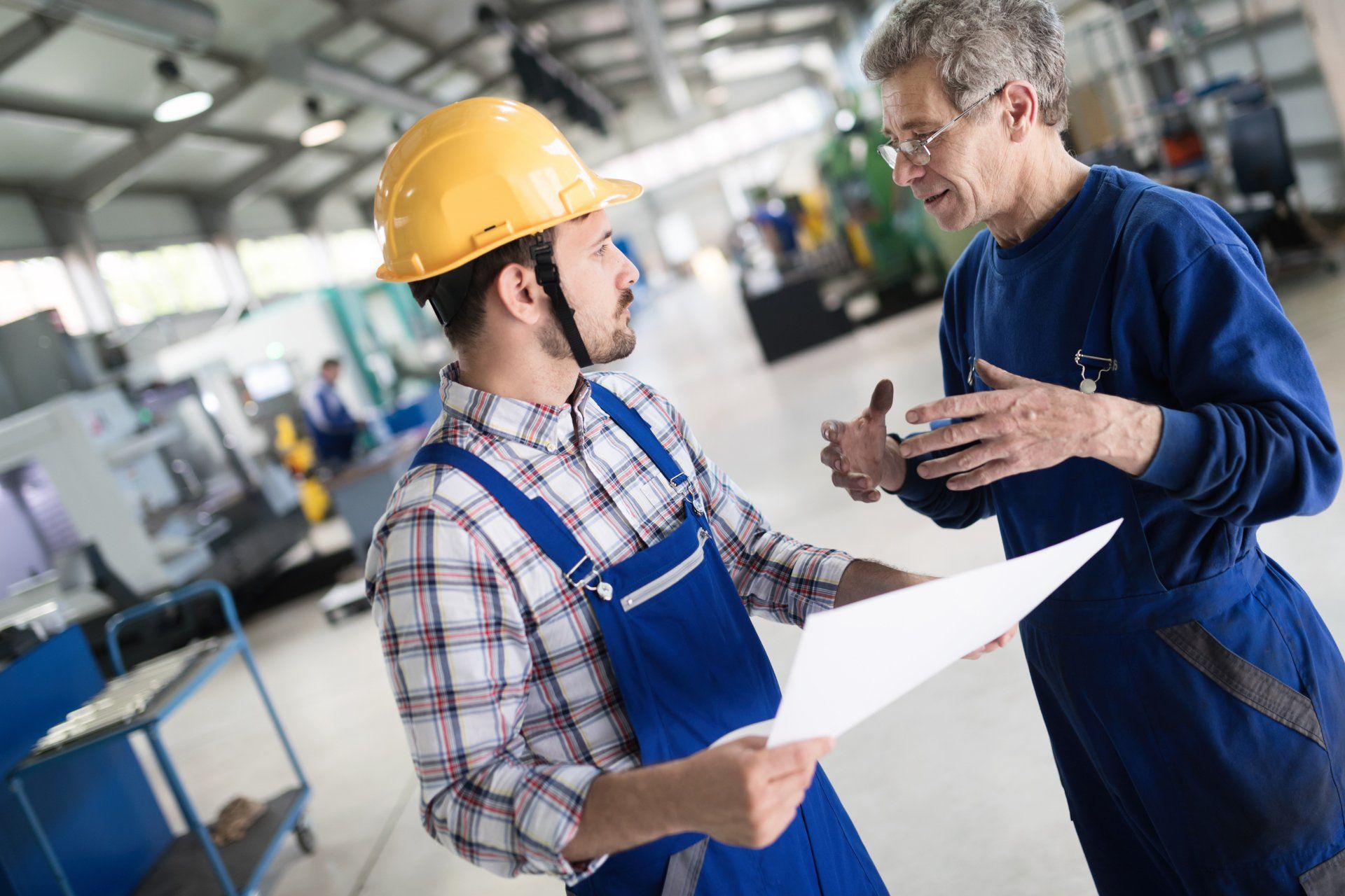 Two men are talking in a factory while one of them is holding a piece of paper.