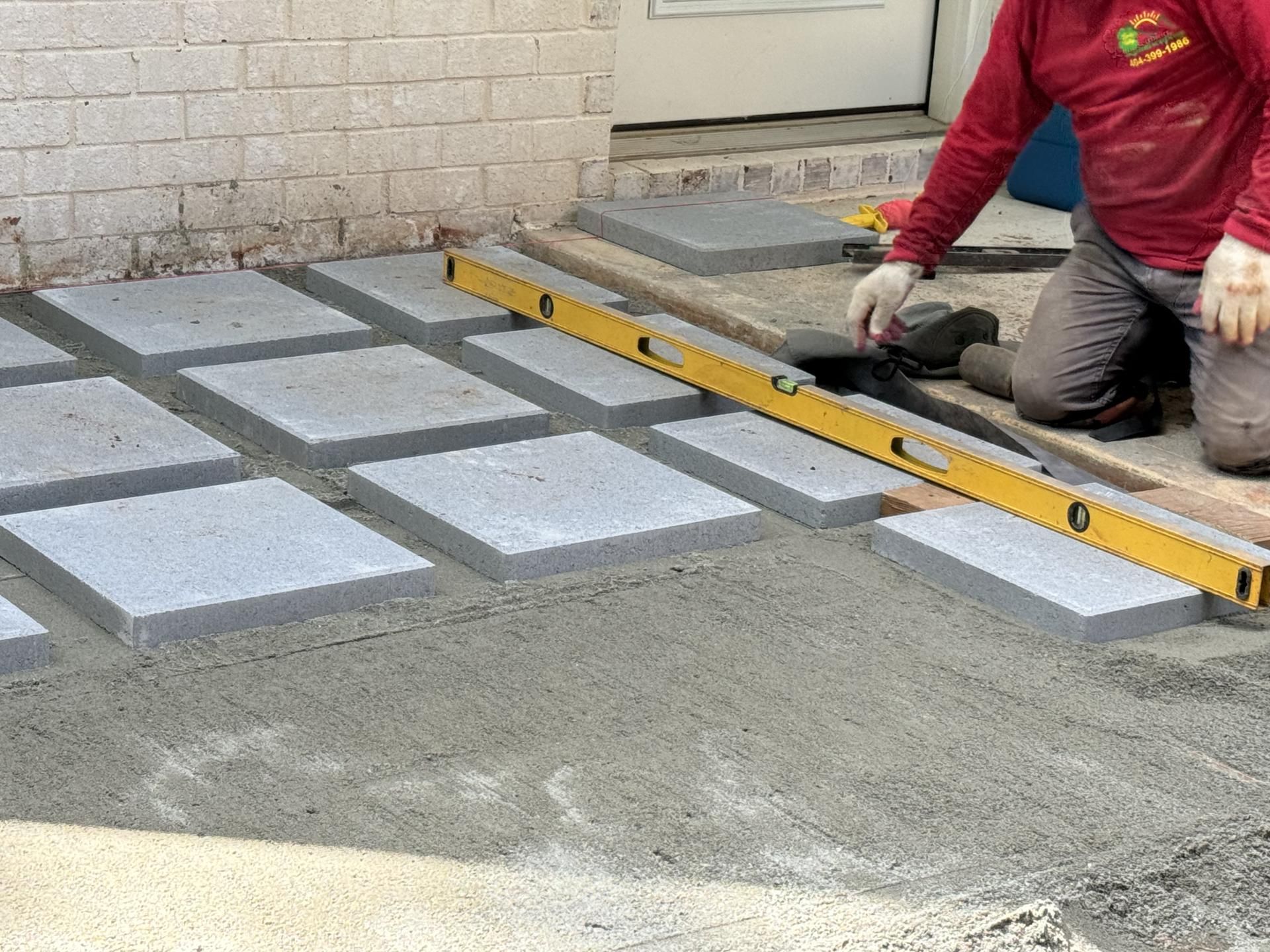 Person laying paving stones with a level in front of a brick wall and a white door.