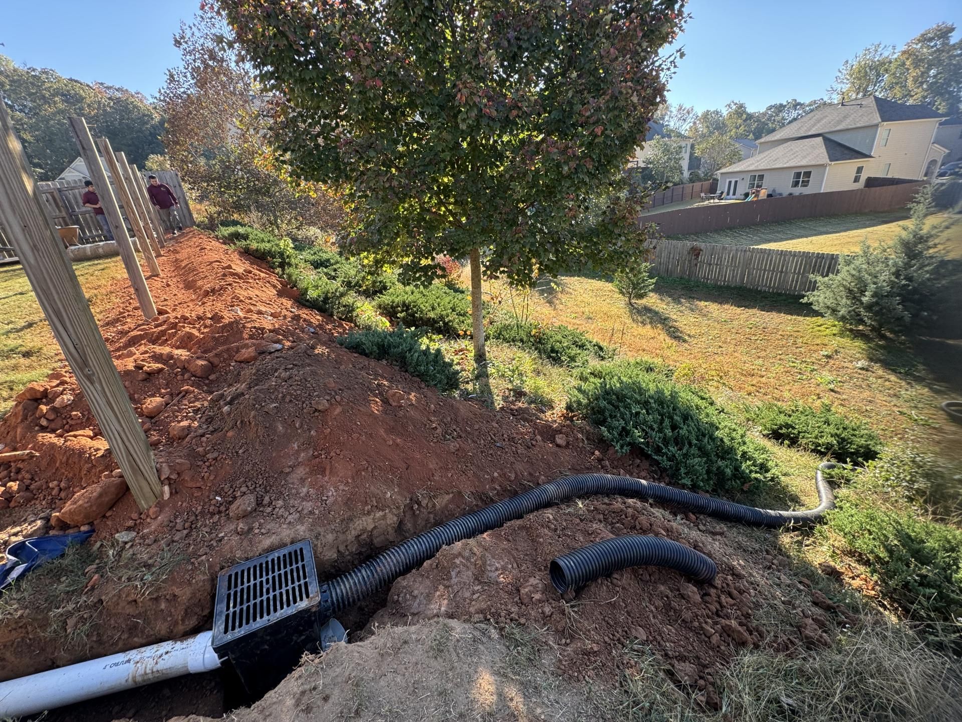 Drainage system installation on a hillside with black pipe and a grate. Brown dirt, green shrubs, and a fence.