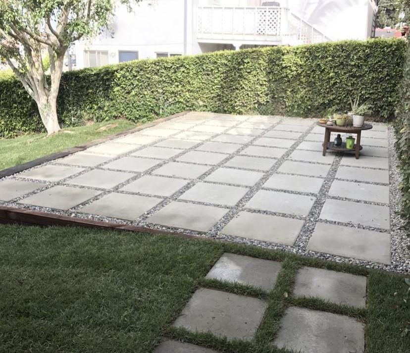 Patio with square concrete tiles, gravel, and grass. A small wooden table holds plants. Green hedge in background.