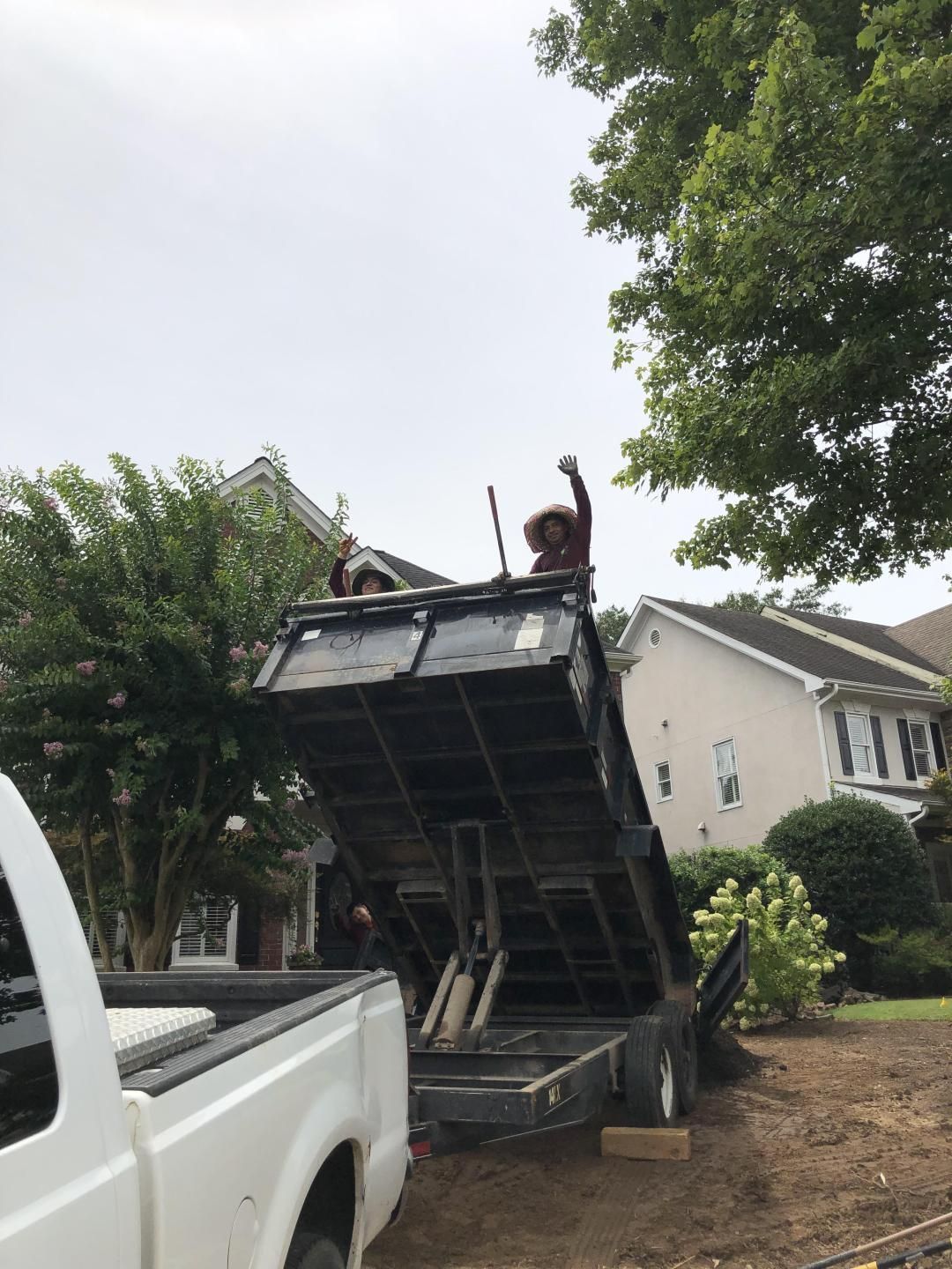 Two people in a raised dump trailer with one arm raised, next to a white truck and houses on a cloudy day.