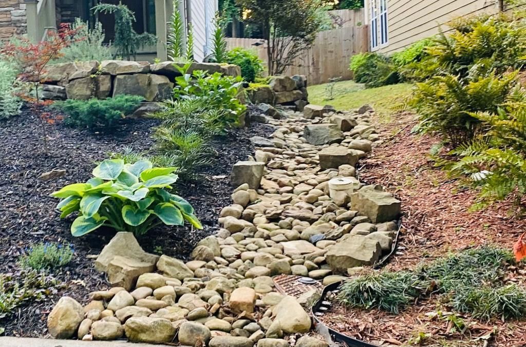 A dry creek bed of stones flows through a landscaped yard with various plants and rock borders.