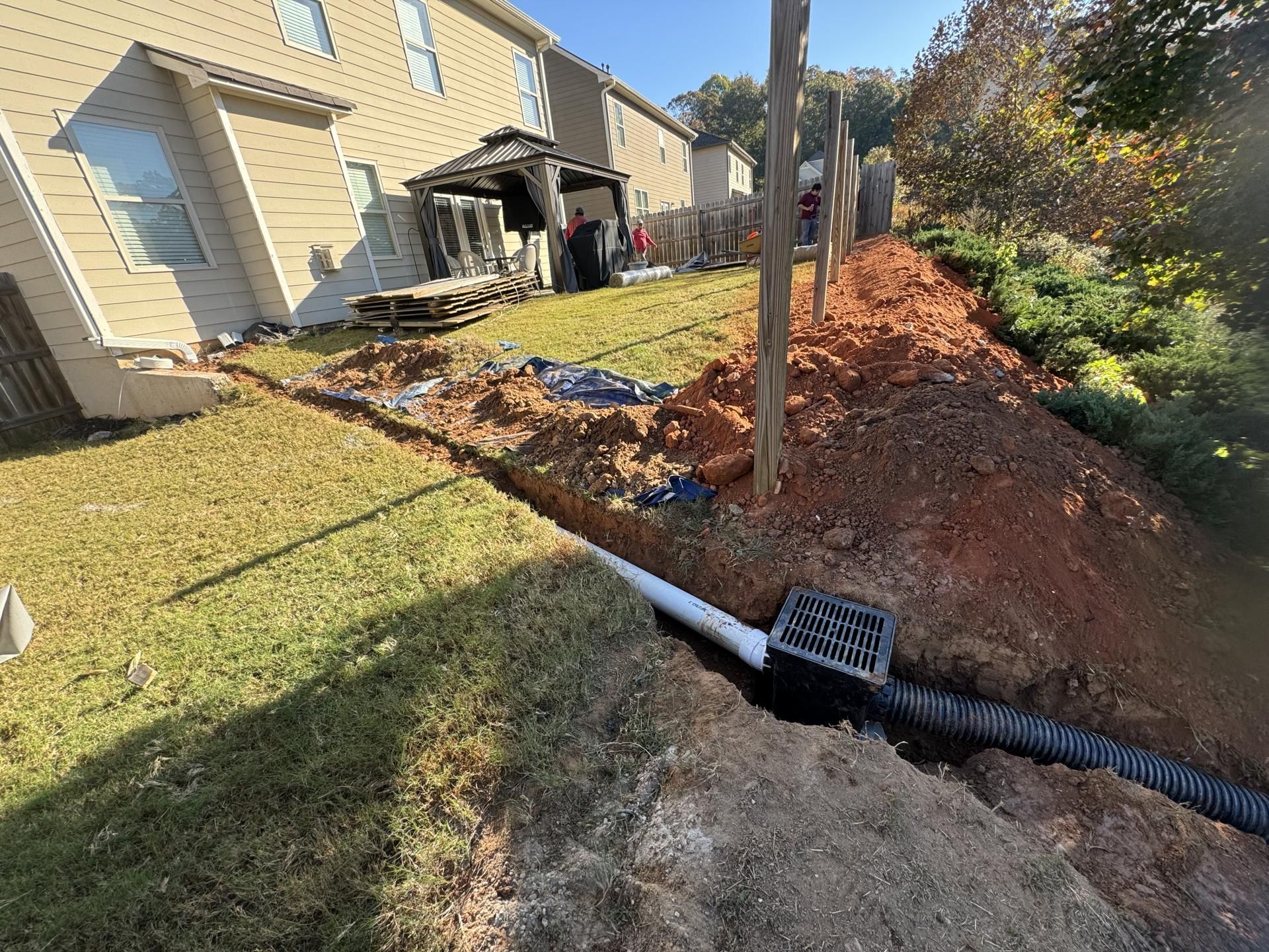 Drainage system installation: ditch with pipe, grated drain, red soil, near houses.