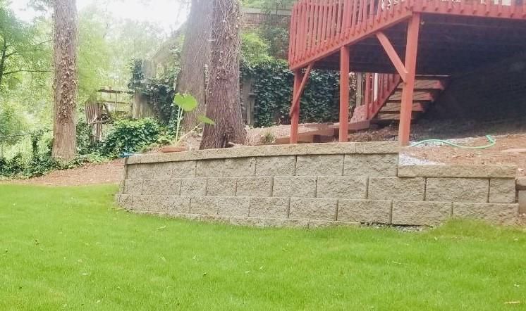 Retaining wall made of gray blocks, supporting a red deck structure; green lawn, trees, and foliage in the background.