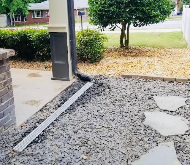 Black downspout drains onto a metal runoff, leading to a gravel bed.