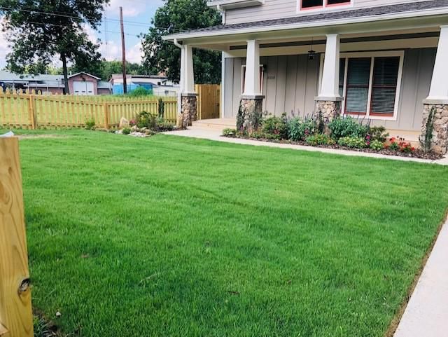 Green lawn in front of a house with a porch and stone columns. A wooden fence lines the yard.