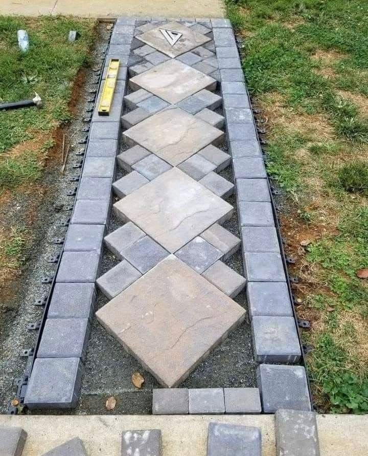 Pathway being constructed with diamond-patterned pavers, bordered by square blocks on gravel, in a grassy setting.
