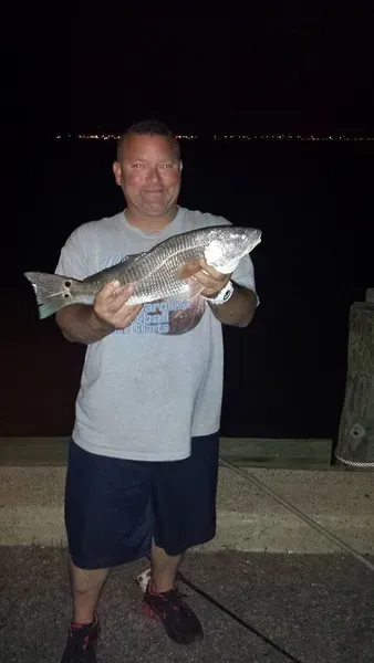 Man holding a large redfish at night on a pier. He is wearing a grey shirt and blue shorts.