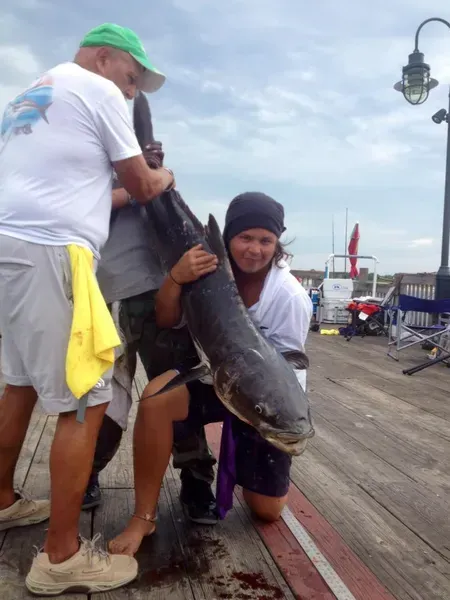 Two people holding a large fish on a wooden dock. The woman kneels and smiles, with the fish in her arms.