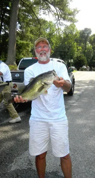 Man in white shirt and shorts holding up a large bass fish near a parking area, smiling.