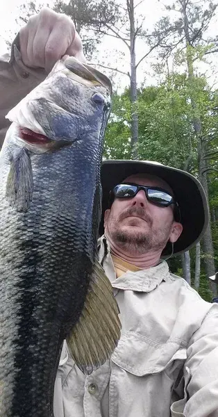 Man holding up a large bass he caught outdoors in front of trees.