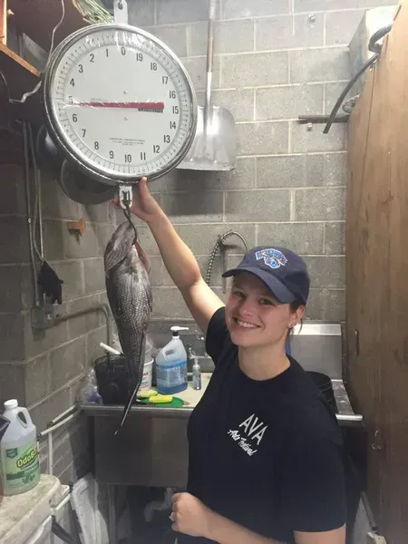 Woman weighing a fish on a scale, smiling in a fish market.