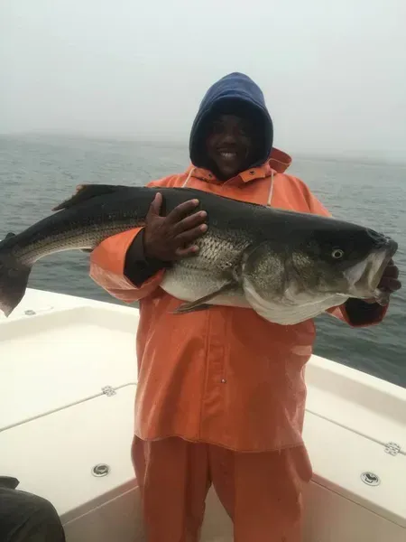Person in orange rain gear holding large fish on a boat, smiling. Gray sky and water in the background.