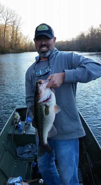 Man in a boat holding a largemouth bass, smiles. Outdoors on a lake, cloudy sky, wearing a hat and gray hoodie.