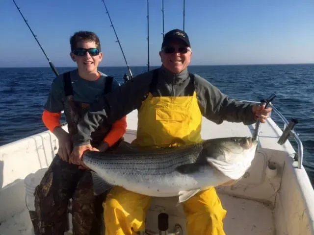 A man and a boy on a boat hold up a large striped bass, blue sea, sunny day.
