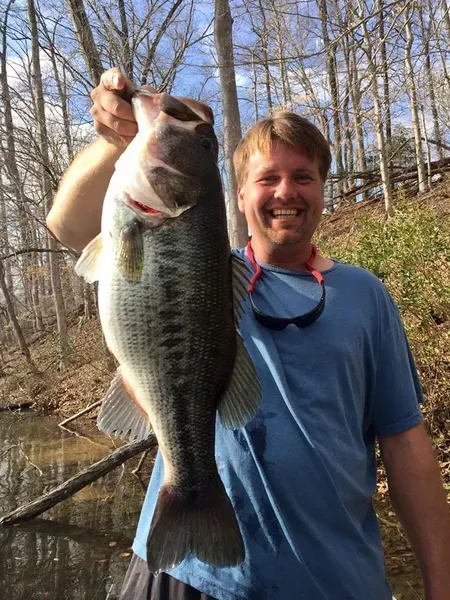 Man smiling, holding a large bass. He wears a blue shirt, sunglasses around his neck, in a wooded area near water.