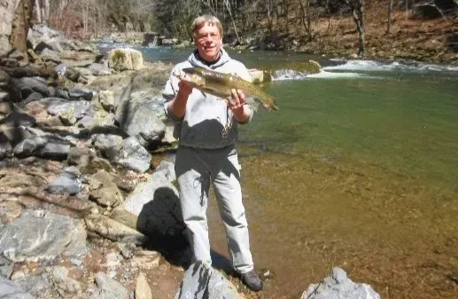 Man holding a fish near a river, wearing a gray hoodie and pants; outdoors.