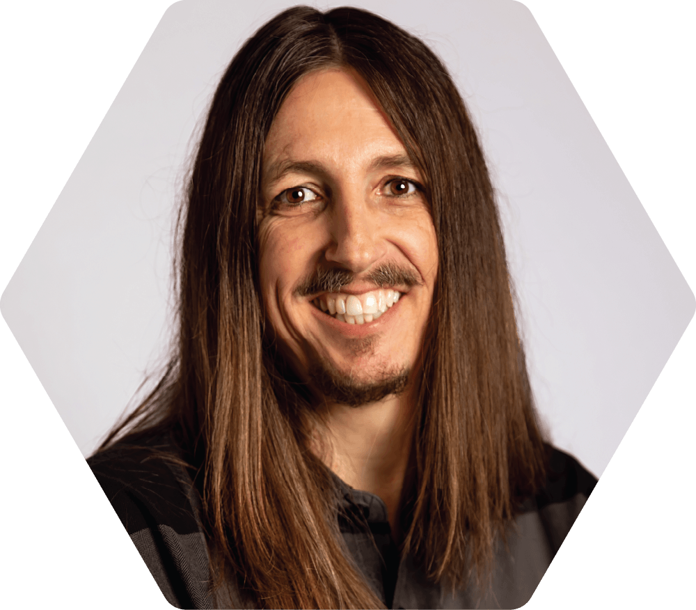 Brian Ricks smiling person with long brown hair in a hexagonal frame, against a white background.
