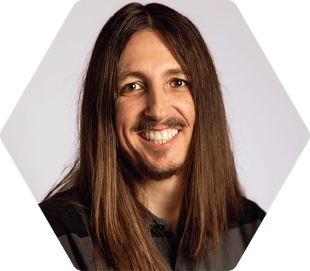 Brian Ricks smiling person with long brown hair in a hexagonal frame, against a white background.