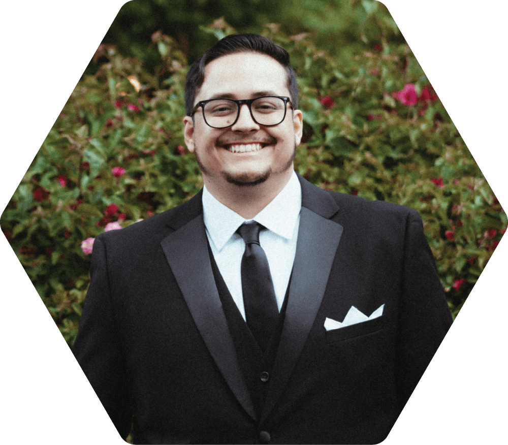 Kobe Ricks in a black tuxedo smiling, wearing glasses, in front of greenery.