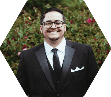 Kobe Ricks in a black tuxedo smiling, wearing glasses, in front of greenery.